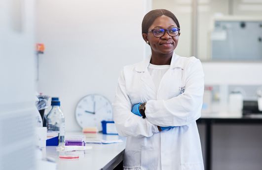 A Black laboratory medicine professionals stands in a lab with her arms crossed, looking thoughtfully into the distance.