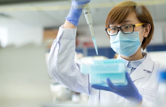 An Asian laboratory medicine professional wearing a mask holds a pipet up in front of her face.