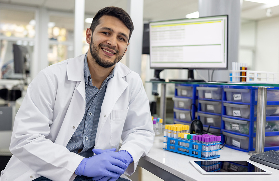 A Latino laboratory medicine professional leans against his desk in a lab.