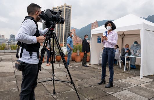 A TV journalist wearing a mask speaks in front of a camera in the midst of a health fair.
