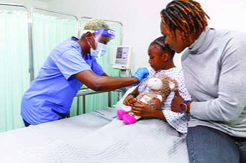 A young girl on a hospital bed with her mother by her side as a female nurse touches her arm.