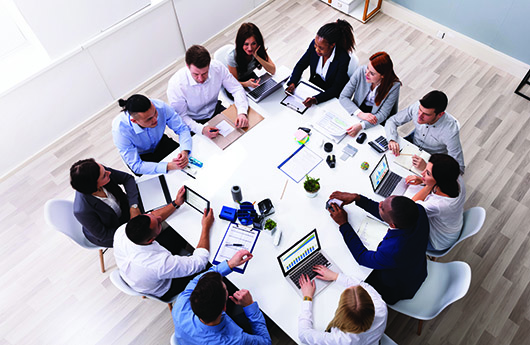 A diverse group of professionals sit around a conference table in a modern office space.