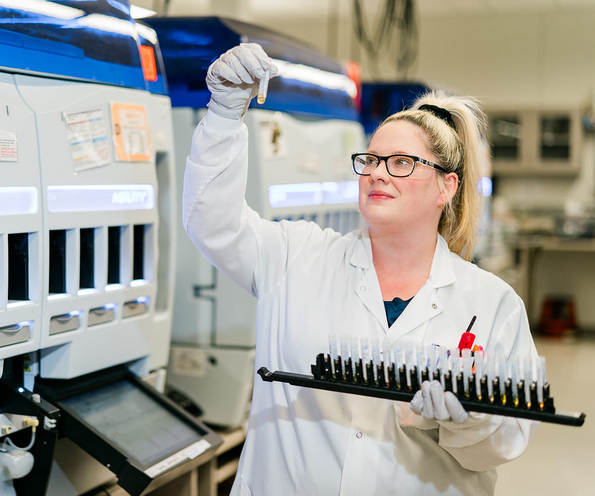 A female laboratorian in a lab, holding a sample tray in one hand and looking at an individiual sample in another hand
