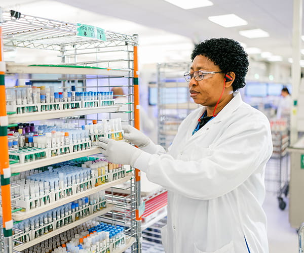 A female laboratory medicine professional adds a rack of tubes to a large shelf full of tubes.