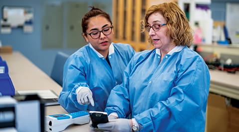 Two female laboratorians in blue coveralls looking at somthing on a bench