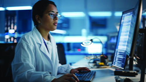 A female laboratorian in a lab, sitting at a desk, and working on a computer