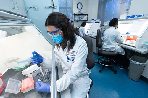 A female laboratorian in a mask holding a pipette in one hand and a sample in the other hand.
