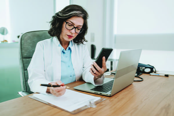 Woman wearing white lab coat works at a desk using a laptop, hand-held device, and paper and pen.