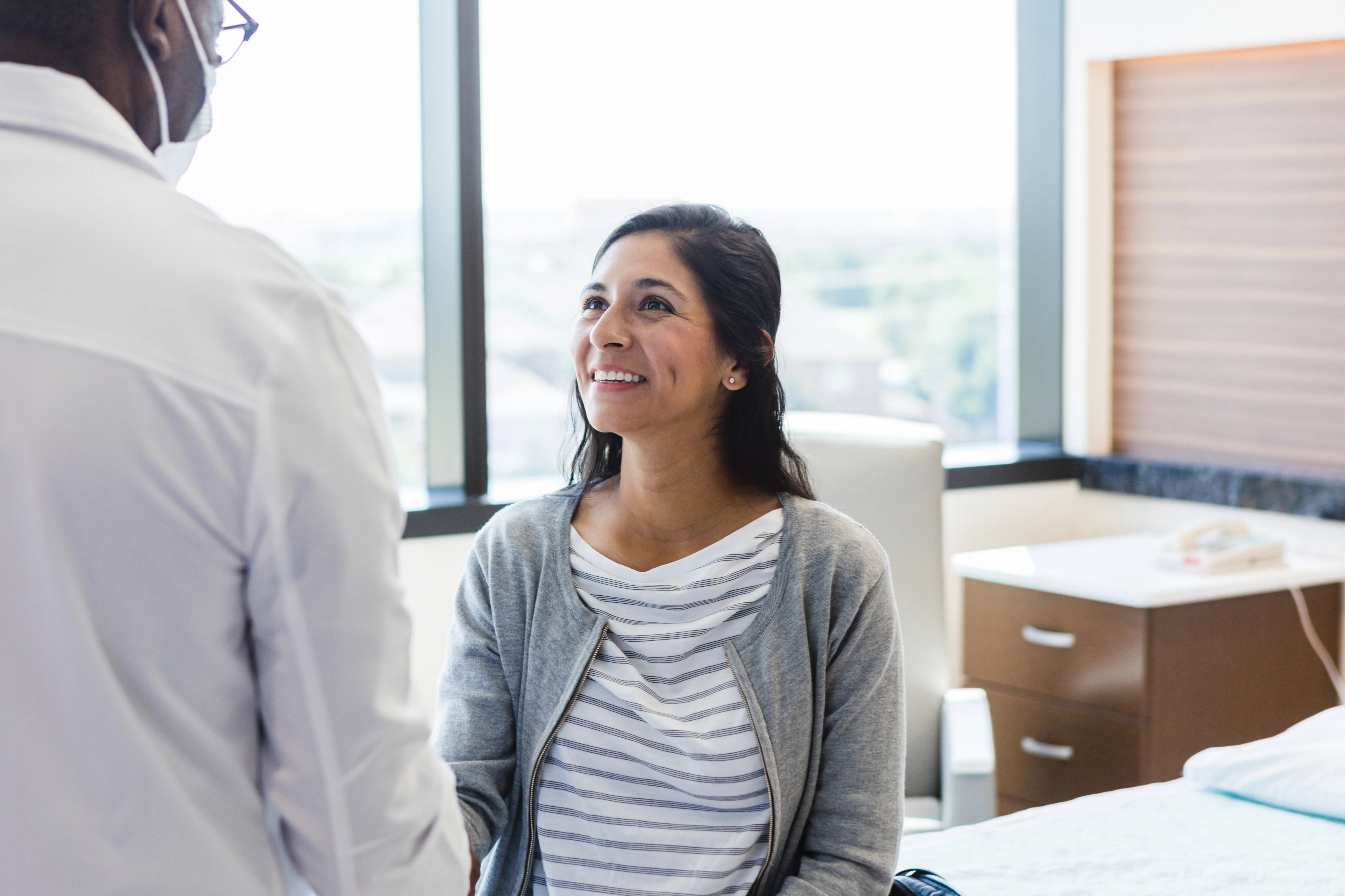 A young adult woman looks up and smiles as the unrecognizable male doctor greets her when he comes in to ER room.