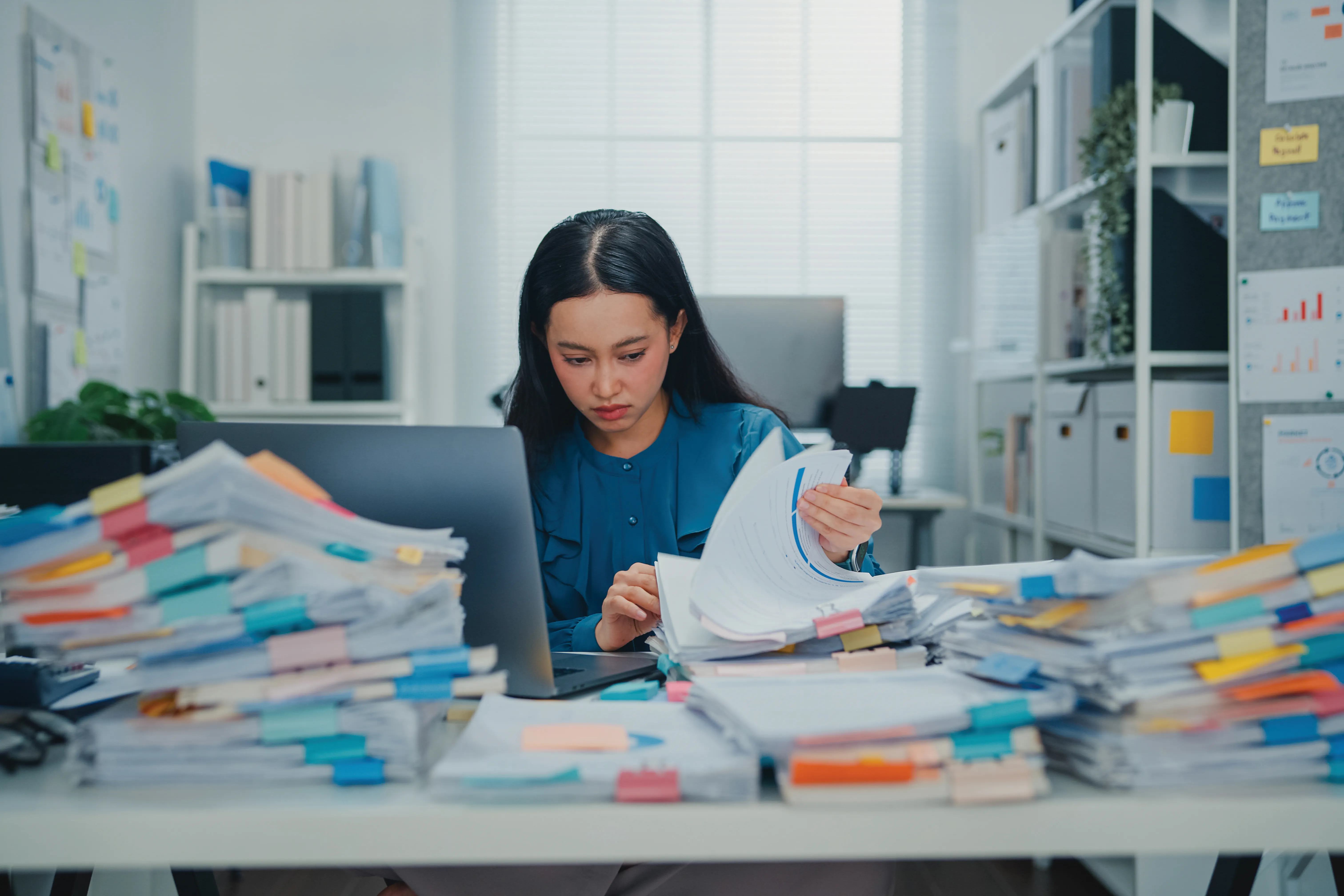 Overworked Asian businesswoman surrounded by piles of documents while working on laptop in busy office. Representing stress, workload, modern corporate life concept.