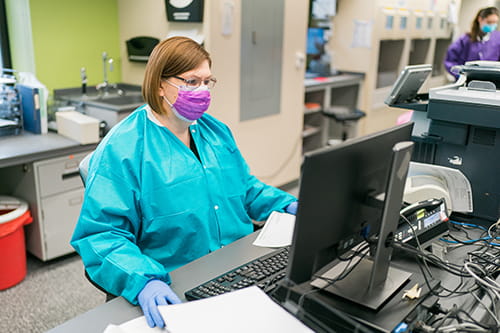 A woman laboratorian in a mask and lab coat sitting at a computer.
