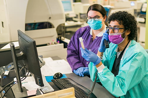 A male and woman laboratorians sitting at a computer, on the phone, while looking at a test tube.