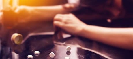 Pills lay scattered on a counter; a personâ€™s arms partially blurred in the background also rests on the counter