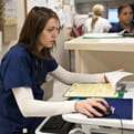 A female healthcare professional with one hand holding an open patient record clicks a computer mouse in the other