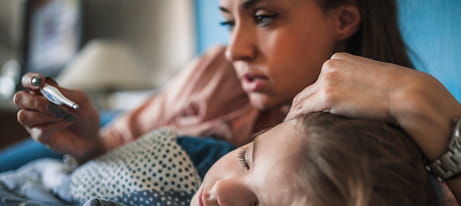 A mother lays beside her child, with one hand on the childâ€™s forehead and with the other holding a thermometer