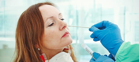 A woman with a mask around her neck is having a swab inserted into her nose by a healthcare provider with gloved hands