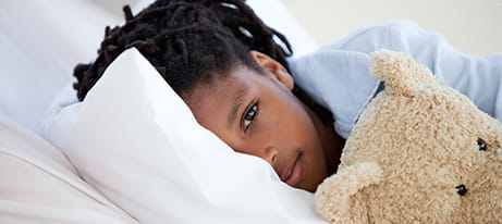 A young girl laying down with half her face obscured by a pillow and holding a teddy bear looks straight ahead