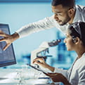 Male and female healthcare professionals, the woman sitting down and the man standing and leaning over, look at a screen the man is pointing to