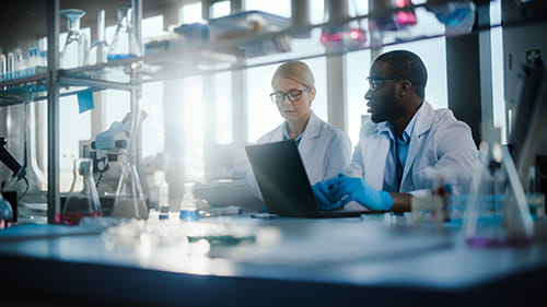 A woman and a man in lab coats, goggles, and gloves, sitting at a desk looking at a notebook and a laptop and lab equpiment around them.